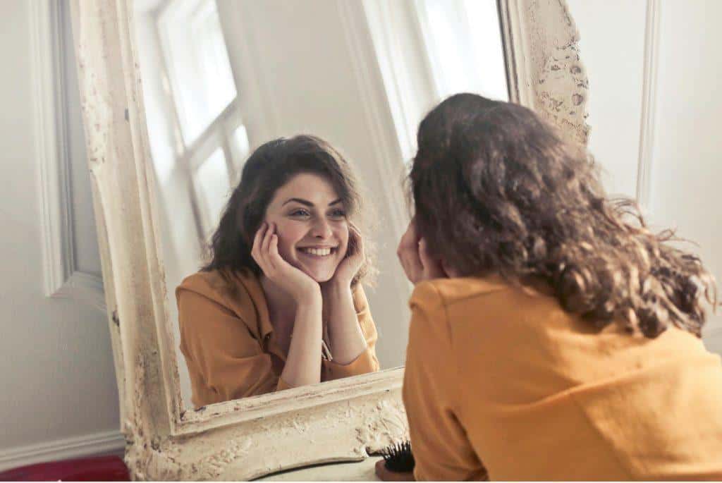 New Year's Resolutions from Tampa Counseling Place in Tampa, Florida: Person in a tan sweater smiling while looking at their reflection in a vintage-style mirror with white ornate frame