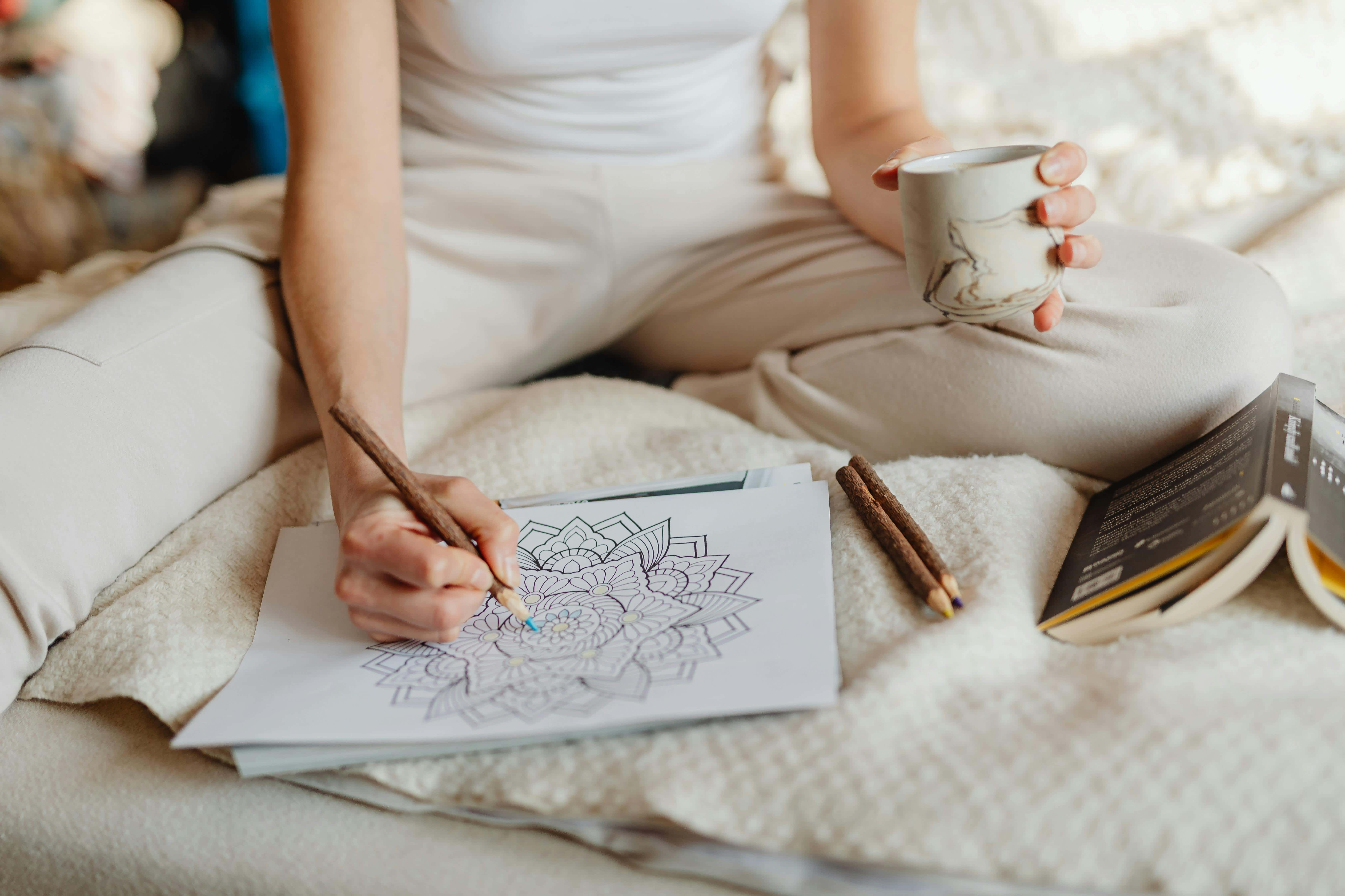 Self-care, Tampa, Fl. at Tampa Counseling Place. This image depicts a person sitting on a soft, light-colored surface, such as a bed or couch, engaged in coloring a mandala design on paper with a wooden pencil. They are holding a ceramic mug in one hand, possibly containing a beverage, and there is an open book and additional pencils placed nearby. 