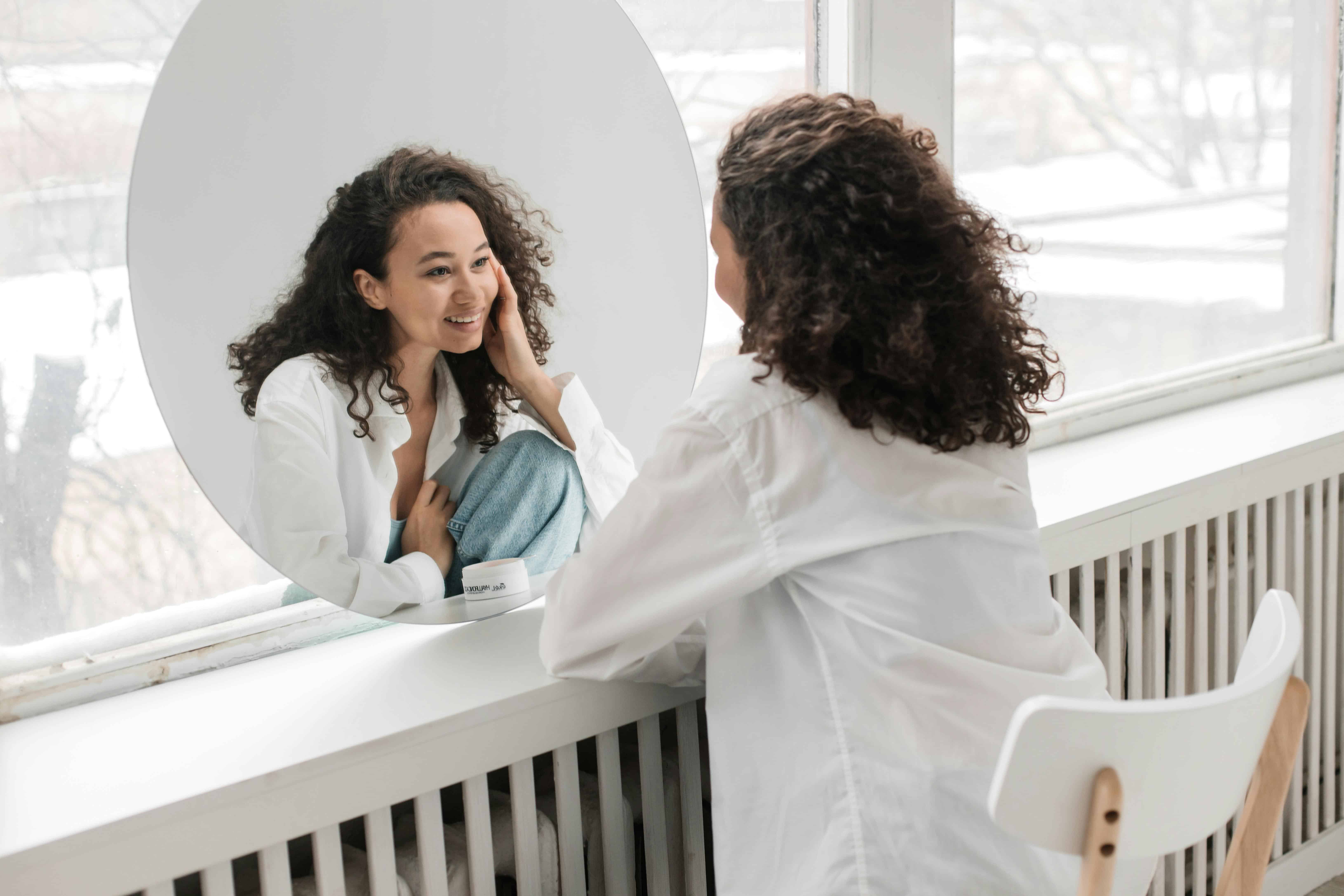 Self-care, Tampa, Fl. at Tampa Counseling Place. This image shows a person with curly hair sitting in front of a circular mirror, smiling and touching their face. They are dressed in a white shirt and jeans, leaning on a white surface, possibly a vanity or windowsill. The setting includes a bright window in the background with snowy scenery visible outside, creating a natural and serene atmosphere.